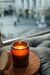 Burning candle in amber glass jar with wooden lid on a cork coaster, placed on a soft grey cushion...