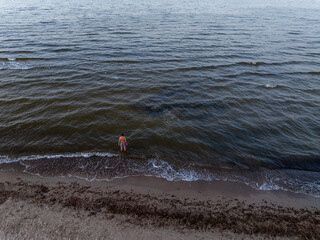 A solitary figure stands at the edge of a beach with dark sand and scattered seaweed, facing gentle...