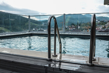 Modern rooftop pool with stainless steel ladder and glass fence. Water reflects cityscape and...