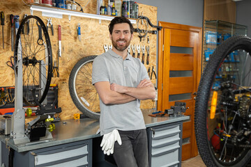 Mechanic standing with crossed arms in bicycle repair shop surrounded by tools and bike parts