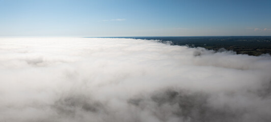 A wide view of dense clouds covering the lower frame, contrasting with a clear blue sky above. The horizon shows a forested area in the distance.