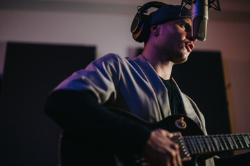 A focused musician in a recording studio plays electric guitar and sings into a microphone. The...