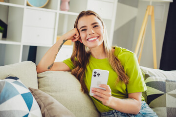 Smiling young woman with long hair seated indoors holding smartphone wearing green shirt in a cozy modern living room