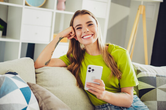 Smiling young woman with long hair seated indoors holding smartphone wearing green shirt in a cozy modern living room