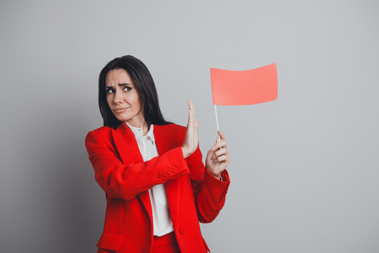Businesswoman in a red suit holding a red flag while making a gesture against the concept, on a neutral grey background