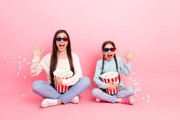 Mother and daughter sitting on a pink background, enjoying 3D movie with popcorn, laughing happily...