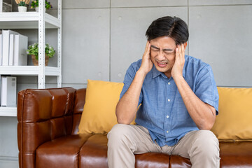 Asian elderly man suffering from migraine headache, holding head in pain while sitting on sofa