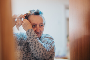 An elderly woman arranges her hair with curlers, wearing a comfortable, patterned sweater. She is...
