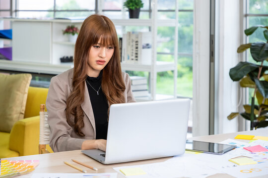 Young Asian woman working on laptop looking frustrated at creative office desk with color swatches and design sketches