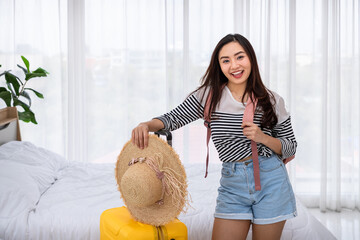 Cheerful young Asian female traveler in striped shirt holding straw hat and suitcase in bedroom...