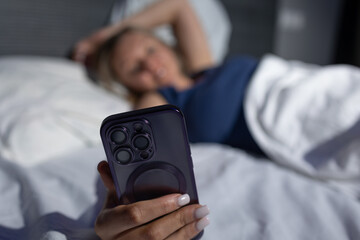 A person is comfortably lying in bed with white bedding, casually checking their smartphone in low light. The setting evokes a peaceful and relaxed nighttime atmosphere, perfect for winding down.