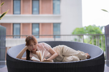 Young Asian woman lying comfortably on a round chair on a terrace, deeply immersed in a book,...