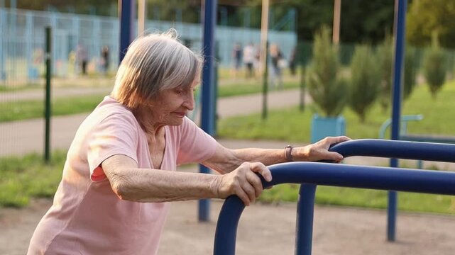 An elderly woman over 80 years old performs a shoulder stretch on an outdoor fitness station. This time, she is turned slightly to the side, adding a gentle twist to the movement. 