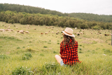 A woman in a red plaid shirt and straw hat sits in a field of grass