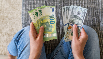A person sitting indoors in jeans while holding and comparing euro and dollar banknotes,...