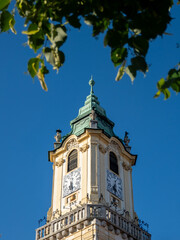 Clock tower of the Jesuit Church in Bratislava, Slovakia, captured on a sunny day framed by green leaves against a vibrant blue sky.