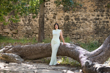 Young, pretty, brunette woman in a white dress, posing looking at camera leaning on the trunk of a...
