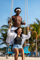 Happy interracial couple enjoying a fun beach day, swinging under palm trees on a tropical vacation