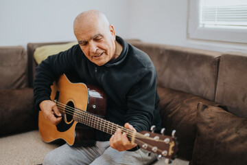 An elderly man sitting on a sofa is playing an acoustic guitar. The setting conveys leisure,...