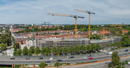 Panoramic view of large urban construction site with four cranes, scaffolding-covered buildings, residential homes in the background and road with traffic in front. Construction site in Kiel, Germany.
