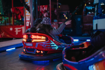 Two individuals laugh and wave as they ride vibrant bumper cars during a joyful evening outing at a nighttime amusement park, illuminated by colorful lights, creating a lively and vibrant atmosphere.