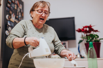 Elderly woman actively mixing food ingredients in the kitchen using an electric mixer,...