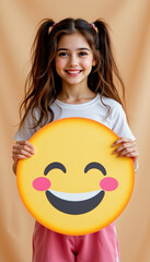 Smiling girl with pigtails holding large happy emoji face with blushing cheeks and closed eyes, cheerful expression on beige background