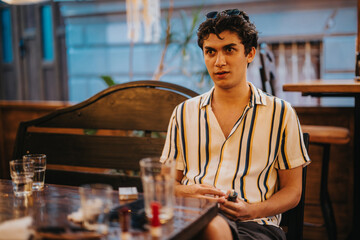 A young man in a striped shirt is seated at a cafe table, exuding a relaxed and stylish atmosphere....