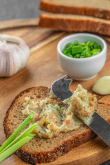A slice of rye bread is generously spread with garlic lard spread and garnished with green onions, surrounded by fresh ingredients on a wooden board