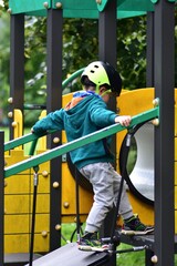 Young boy climbing modern playground bridge, wearing safety helmet and green hoodie. Action shot capturing balance, focus, and confidence during outdoor play, promoting healthy childhood development.
