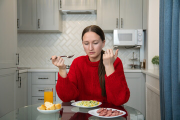 Young woman sitting at kitchen table, holding fork and looking at plate of salad and plate of...