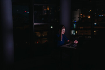 A woman sits in a dark office, illuminated by her laptop screen, appearing stressed. The city...