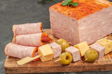 Rolled ham slices, cheese cubes, olives, and skewered appetizers are arranged on a wooden board next to a large block of ham loaf for a hearty and inviting snack display