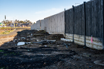 An urban fringe behind a timber fence, strewn with construction debris, rubbish, and overgrown...