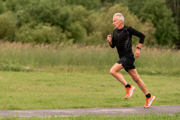An elderly man running on a paved path in a green park.