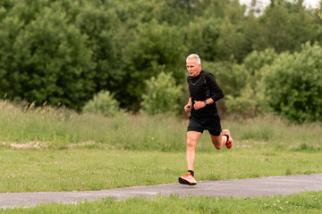 An elderly man running on a paved path in a green park.
