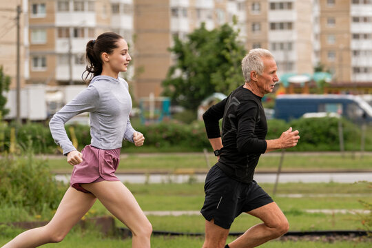 elderly man and a young girl run along a path in a city park together.