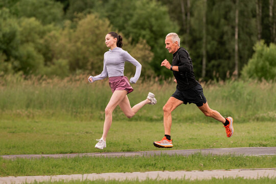 elderly man and a young girl run along a path in a city park together.