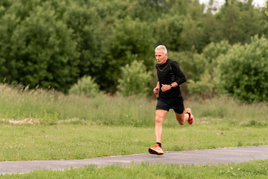 An elderly man running on a paved path in a green park.
