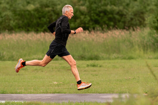 An elderly man running on a paved path in a green park.