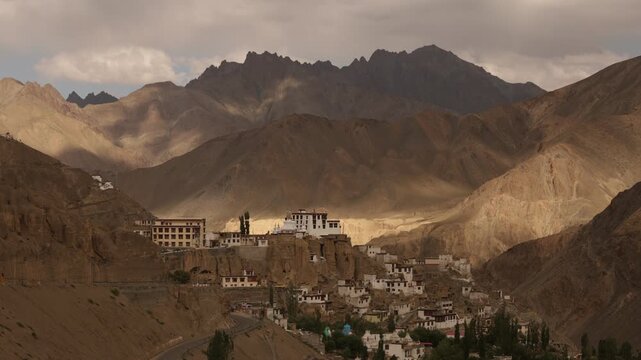 Gompa nestled on cliff with village below, dramatic shadow and light from moving clouds in mountainous valley landscape.