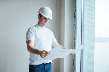 A young man carefully studies a blueprint, standing by a window. A foreman in a white helmet...