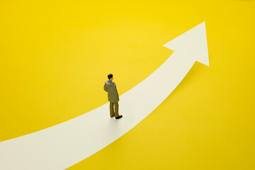Concept of challenge to success and growth of business career path. Businessman doll walks along a path with arrow pointing forward on yellow background.