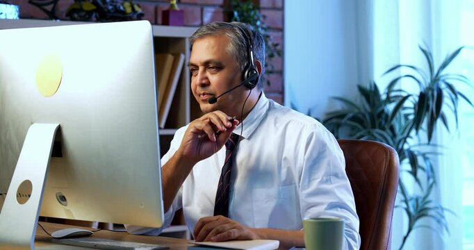 Indian call center executive in stress with overload, sitting at desk in modern office wearing headphones, Asian corporate customer service employee attending nonstop calls on computer system