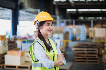 Portrait of smiling Southeast Asian female warehouse worker in safety gear standing confidently with arms crossed inside storage facility.