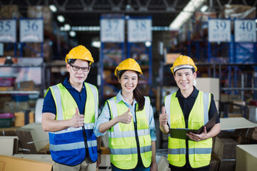 Group of Southeast Asian warehouse employees wearing safety helmets and reflective vests giving thumbs-up gesture, standing in storage area with stacked boxes in background.