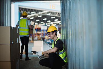 Logistics workers loading cardboard boxes into shipping container, wearing safety helmets and vests, in busy warehouse with open dock.