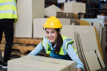 Smiling Asian female warehouse worker wearing yellow hard hat and reflective vest lifts cardboard...