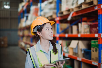 Asian female warehouse worker wearing safety vest and helmet uses digital tablet to manage...