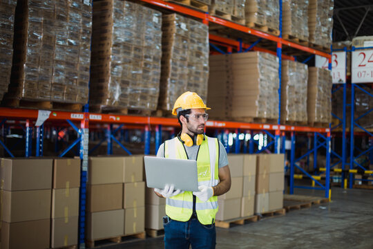 Warehouse logistics coordinator wearing safety gear using laptop to manage transportation schedules and cargo operations in a busy industrial distribution center.